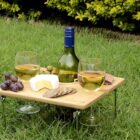 A small wooden picnic table sits on grass, holding a bottle of white wine, two glasses filled with wine, green olives, red grapes, a wedge of cheese, and crackers. Bushes are visible in the background.