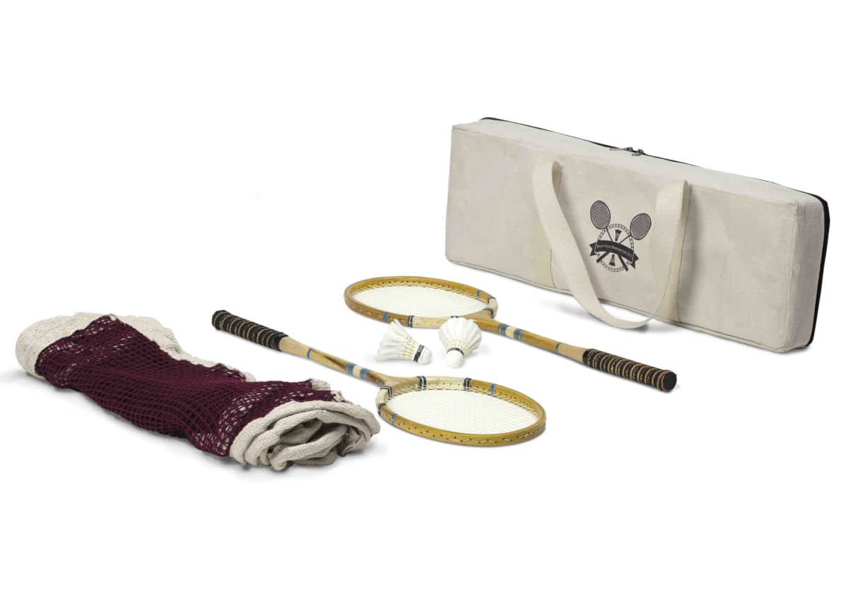 A vintage badminton set with two rackets, shuttlecocks, a net, and a carrying case. The rackets have wooden frames and the net is partially rolled up beside them. The carrying case is beige with a logo featuring two shuttlecocks.
