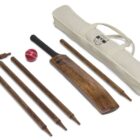A cricket set displayed on a white surface, including a wooden cricket bat, a red cricket ball, wooden stumps with bails, and a beige carrying bag.