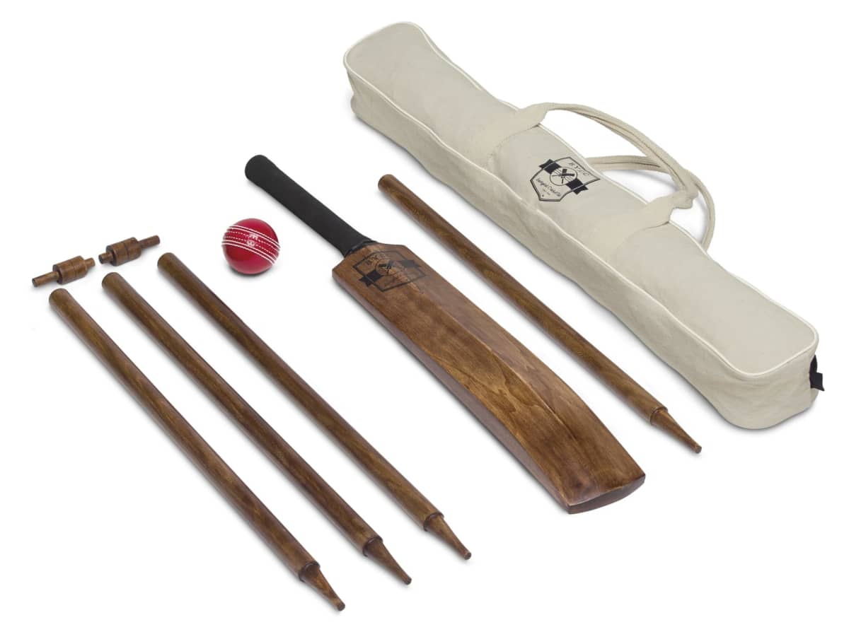 A cricket set displayed on a white surface, including a wooden cricket bat, a red cricket ball, wooden stumps with bails, and a beige carrying bag.