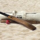A wooden cricket bat, a red cricket ball, and three wooden stumps are arranged on sandy ground. Behind them is a beige, cylindrical cricket kit bag with BYCC printed on it, featuring crossed bats and a crown.
