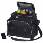 A black cooler bag with built-in speakers, containing apples, an orange, a bottle of water, and a bottle of wine. An MP3 player is connected to the speakers via a cable. The bag has a shoulder strap and unzipped top.