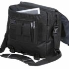 A black messenger bag with multiple compartments and pockets, containing a notepad and a few pens. The bag has a wide shoulder strap and side buckles, with an open flap revealing its contents.