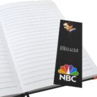 An open lined notebook with a black bookmark featuring a person, the word House, and the NBC logo. The bookmark is placed on the right page.