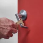 A hand uses a no-touch tool to open a silver door handle on a red door. The tool is designed to reduce direct contact with surfaces, enhancing hygiene. A key is visible in the hands grip.