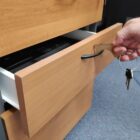 A hand uses a key to unlock a drawer in a wooden filing cabinet. The drawer is partially open, revealing a black binder inside. The cabinet is located in a room with a carpeted floor.