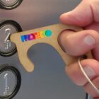 A hand uses a key-shaped tool to press the button for the second floor in an elevator. The tool is brown with the word PROTECO in colorful letters.