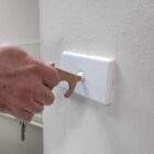 A hand uses a metal key-like tool to press a light switch on a white wall.