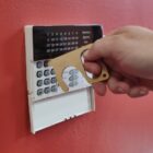 Hand using a brass touch tool to press a button on a wall-mounted keypad security system. The panel is white, and the background wall is red.