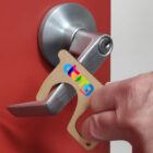 A person uses a handheld tool to open a door with a silver handle. The tool is shaped like a key and is made of a beige material with a colorful logo on it. The door is red.