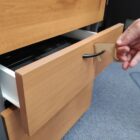 A hand using a small tool to pull open a wooden drawer by its handle. The tool allows the user to open the drawer without direct hand contact. The drawer is part of a light wood cabinet on a carpeted floor.