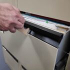 A hand opening a beige filing cabinet drawer, revealing a collection of files and folders inside. The person is holding one of the folders by the tab. The scene is set in an office environment.