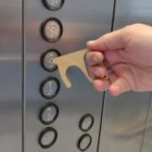 A hand using a brass tool to press a button inside an elevator. The tool helps avoid direct contact with surfaces, promoting hygiene.