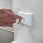 A person uses a brass no-touch tool to press a light switch on a white wall. The tool helps avoid direct contact with surfaces.
