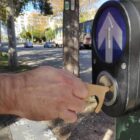 A person uses a tool to press a pedestrian crossing button on a city street. The scene is outdoors with trees and cars visible in the background.
