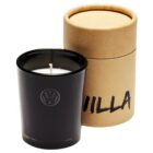A black candle in a glass holder with a logo on the front is placed next to a cylindrical cardboard box labeled VILLA. The box has a matching black logo and a lid partially on. The candle appears to be unused.