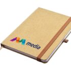 A brown notebook with an elastic band. The cover displays a colorful logo with the letters AM followed by the word media. A ribbon bookmark is visible at the edge of the notebook.