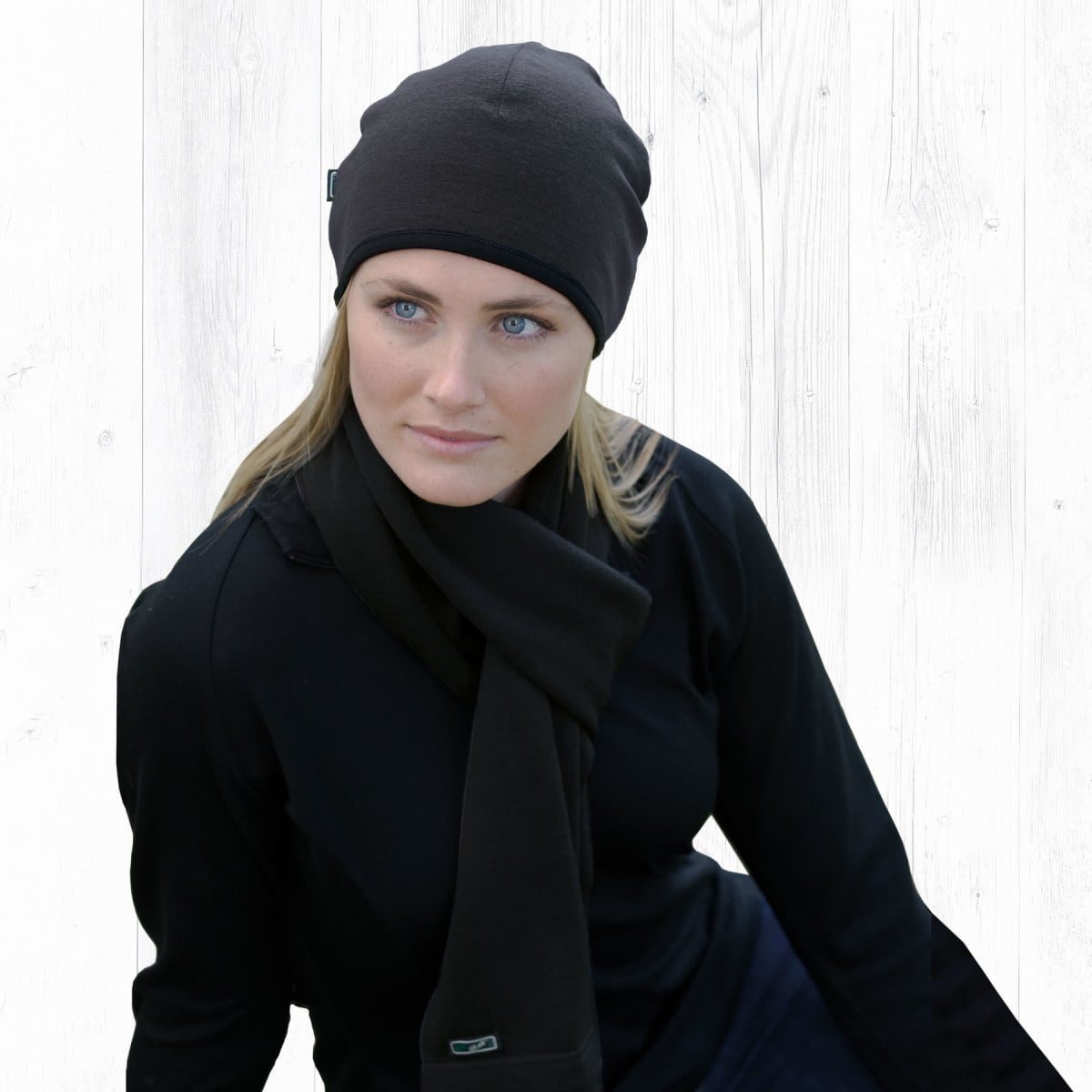 A person with light hair wearing a black beanie, black scarf, and black top sits against a white wood backdrop, gazing thoughtfully to the side.