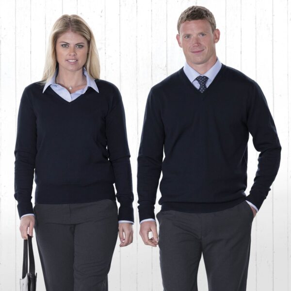 A man and a woman stand side by side against a light background. Both are wearing dark V-neck sweaters over light blue shirts, with gray pants. The woman carries a handbag, and the man has one hand in his pocket.
