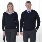 Two people dressed in matching professional attire stand against a plain white background. Both wear navy blue sweaters over light blue collared shirts and dark gray trousers. The person on the left carries a bag.