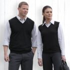 A man and a woman stand side by side in front of a white wooden wall. They are both wearing white collared shirts with black vests. The man has rolled-up sleeves and gray trousers, while the woman holds a bag in her left hand.