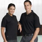 A woman and a man standing together, both wearing black polo shirts and gray pants. They are smiling and posing against a light wood-textured background.