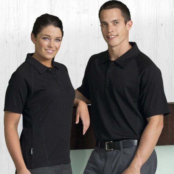 A woman and a man standing together, both wearing black polo shirts and gray pants. They are smiling and posing against a light wood-textured background.