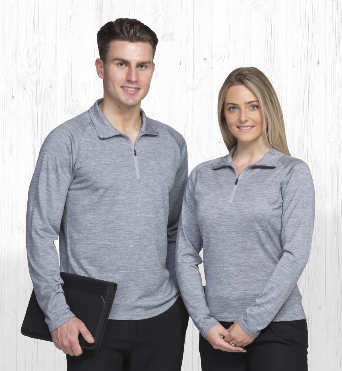 A man and woman stand side by side, both wearing matching gray long-sleeve shirts with half-zip collars. The man holds a black portfolio, and they are smiling against a light wooden background.