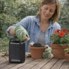 A woman pots flowers outdoors, wearing gloves, with Ellis Outdoor Bluetooth Speakers nearby.