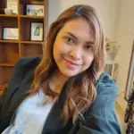 Meet the Team: A person with long hair smiles beside a bookshelf indoors.