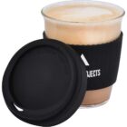 A glass cup filled with a frothy coffee drink, covered with a black silicone sleeve, sits next to a matching black silicone lid. The sleeve has white text and a logo partially visible. The lid reads CAUTION VERY HOT.