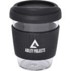 A clear reusable coffee cup with a black silicone lid and grip sleeve. The sleeve features a white triangle logo with the text Ability Projects beneath it.