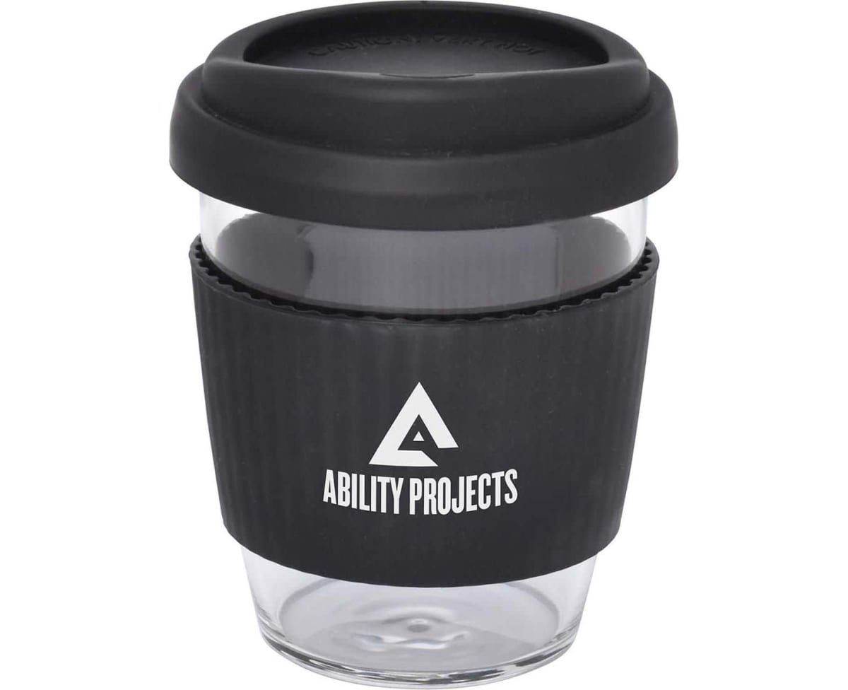 A clear reusable coffee cup with a black silicone lid and grip sleeve. The sleeve features a white triangle logo with the text Ability Projects beneath it.