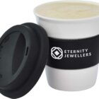A white reusable coffee cup with a black silicone lid and sleeve. The sleeve is printed with Eternity Jewellers and a logo. The cup contains a frothy beverage, and the lid is placed slightly ajar.