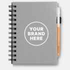 A gray spiral-bound notebook with a circle on the cover that reads YOUR BRAND HERE. An orange pen is attached to the side of the notebook using a black holder.