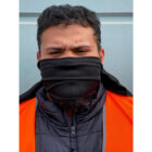 A person with a beard wears an orange vest and a black scarf covering their lower face. They are looking directly at the camera with a neutral expression, standing against a light blue background.