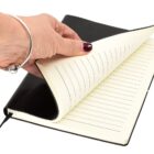 A hand with a silver bracelet is flipping through the lined pages of a black leather notebook. The notebook is open and resting against a white background.