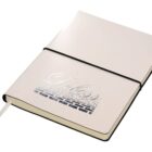 A closed beige notebook with an elastic band and a ribbon bookmark. The cover features embossed text with intricate design elements.