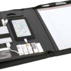 An open black organizer containing a tablet, a smartphone, earphones, a power bank, various cables, batteries, business cards, a pen, and a lined notepad.