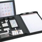 A black zippered portfolio organizer open to display an iPad on the left, a smartphone below it, a power bank, cables, business cards, and a pen. On the right, theres a lined notepad with a pen in the holder.