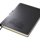 A black leather-bound notebook lies closed, with Deless The Premier Option embossed on the cover. A black ribbon bookmark extends from the bottom. The notebook has rounded edges and a sleek, elegant design.