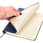 A hand with purple nail polish flips through the pages of a blue, lined notebook. The notebook has a ribbon bookmark and is open on a white background.