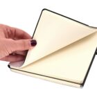 A hand with purple nail polish flips through the blank pages of an open notebook. The notebook has a black cover and plain, cream-colored pages.