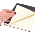 A hand with a bracelet opens a black hardcover notebook, revealing lined pages inside.