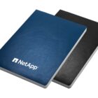 Two closed notebooks stacked slightly askew. The top notebook has a blue cover with the NetApp logo in white, while the bottom notebook is black. Both have white edges, suggesting the presence of lined or blank pages inside.