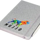 A closed gray notebook with a colorful geometric lion design and the word MUJJO on the cover. A pen is tucked into the notebooks elastic band.
