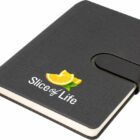 A black notebook with a strap closure, featuring the words Slice of Life and an image of a sliced orange on the cover.