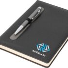 A black notebook with a pen holder integrated into the front cover, holding a silver pen. The cover features a blue and white geometric logo and the text AUSGLASS. A black bookmark ribbon is visible.
