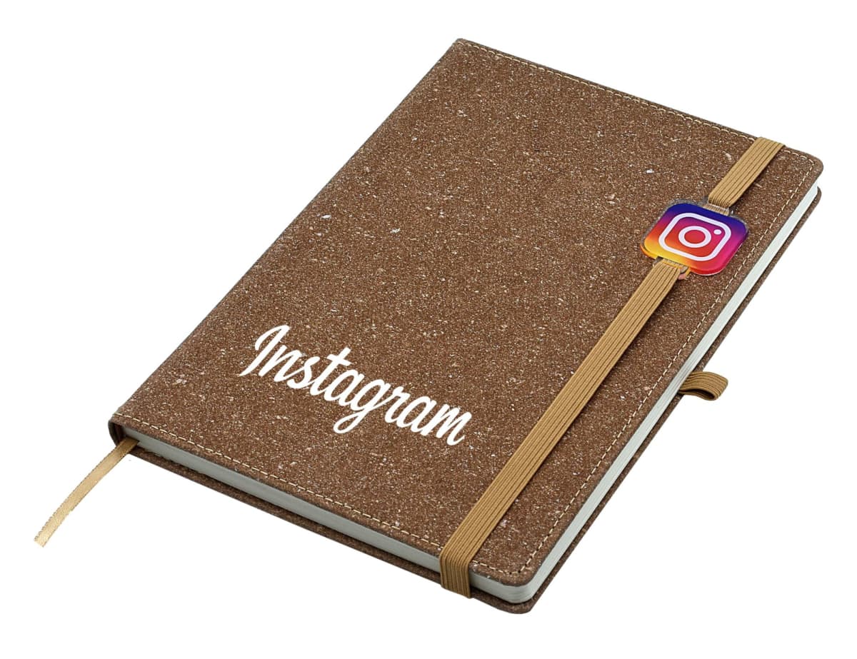 A brown textured notebook with the word Instagram in white on the cover. An elastic band secures it, featuring an Instagram logo charm. The notebook includes a ribbon bookmark.
