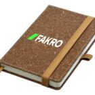 A brown cork-textured notebook with a tan elastic strap is shown. The cover displays the word FAKRO in white with a small green rectangle above the letter A.